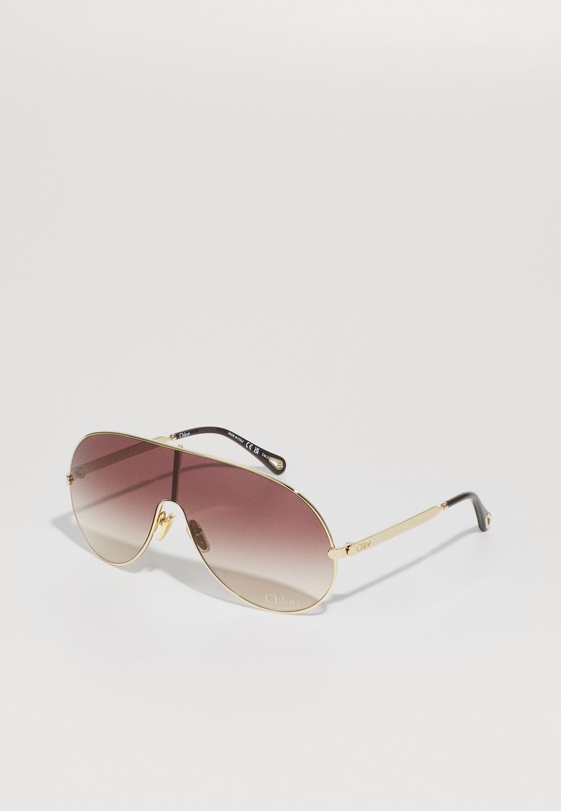 Kuldseid pilootprille, millel on pruunid gradient läätsed, õhuke metallraam ja mustad aksendid templitel, ühel küljel logoga.