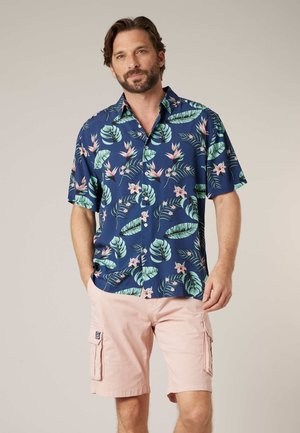 Homme portant une chemise à manches courtes bleu marine avec un imprimé floral tropical rose et vert, et un short cargo rose clair, debout les mains dans les poches.