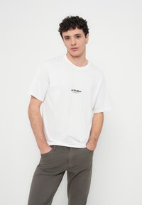 Balti medvilniniai trumpomis rankovėmis marškinėliai su apvalia iškirpte; priekyje juodas „JJ Studios“ logotipas. Derinami su pilkomis kelnėmis.