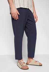 Laivastonsiniset lyhyet housut, jotka on valmistettu sileästä kankaasta, yhdistettynä beigeihin mokkasandaaleihin, joissa on kaksinkertainen solki ja teksturoitu pohjallinen.