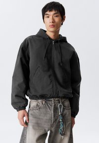 Jonge man in een zwarte rits-jas met capuchon en vervaagde grijze jeans, met een sleutelhanger aan de riemlus geklikt.
