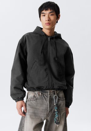 Young man wearing a black zip-up hooded jacket and faded gray jeans with a keychain clipped to the belt loop.