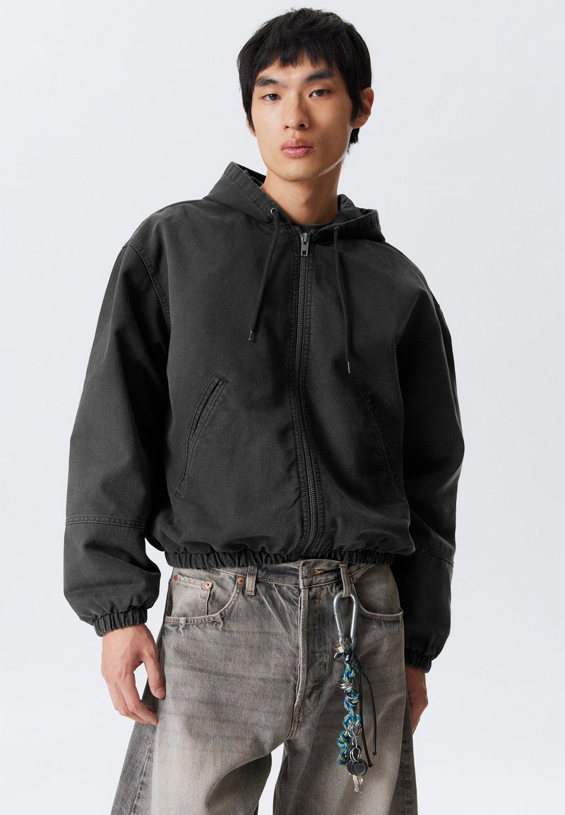 Jonge man in een zwarte rits-jas met capuchon en vervaagde grijze jeans, met een sleutelhanger aan de riemlus geklikt.