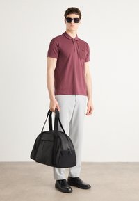 Bordový polo tričko s golierom, svetlošedé nohavice a čierne slnečné okuliare. Drží čiernu športovú tašku s textúrovaným povrchom a logom.