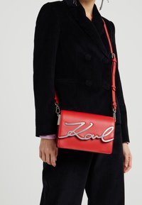 Žena nosící černou kordurovým dvouřadový blazer a kalhoty, nesoucí červenou kabelku přes rameno se stříbrným nápisem "Karl".