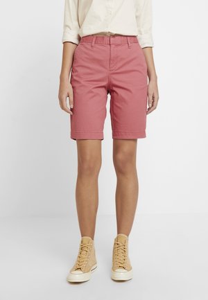 Persoon draagt roze shorts tot halverwege de dij, beige hoge sneakers en een opgerolde witte blouse met lange mouwen, staand tegen een effen achtergrond.