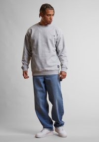 Felpa grigia con polsini e orlo a coste, abbinata a jeans in denim blu dal taglio morbido e sneakers bianche, in piedi su uno sfondo neutro.