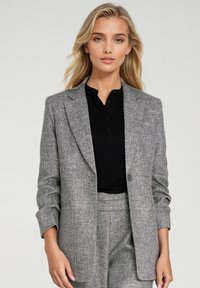 Szary, fakturowany żakiet o dopasowanym kroju, z pojedynczym zapięciem na guzik i wyłóżnymi klapami. Noszony na czarnym topie z kołnierzykiem.