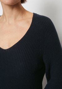 Närbild av en person iförd en mörk marinblå ribbad stickad tröja med djup V-ringning och långa ärmar mot en neutral bakgrund.