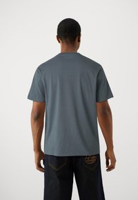 Pelēka kokvilnas T-kreklā ar īsām piedurknēm, noapaļotu kakla izgriezumu un brīvu piegriezumu. Redzami izšuvumi gar šuvēm, valkāta ar tumšām džinsām.