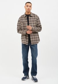 Camisa de cuadros en beige, marrón y azul marino, superpuesta a una camiseta negra. Se lleva con jeans azules y zapatillas deportivas grises.