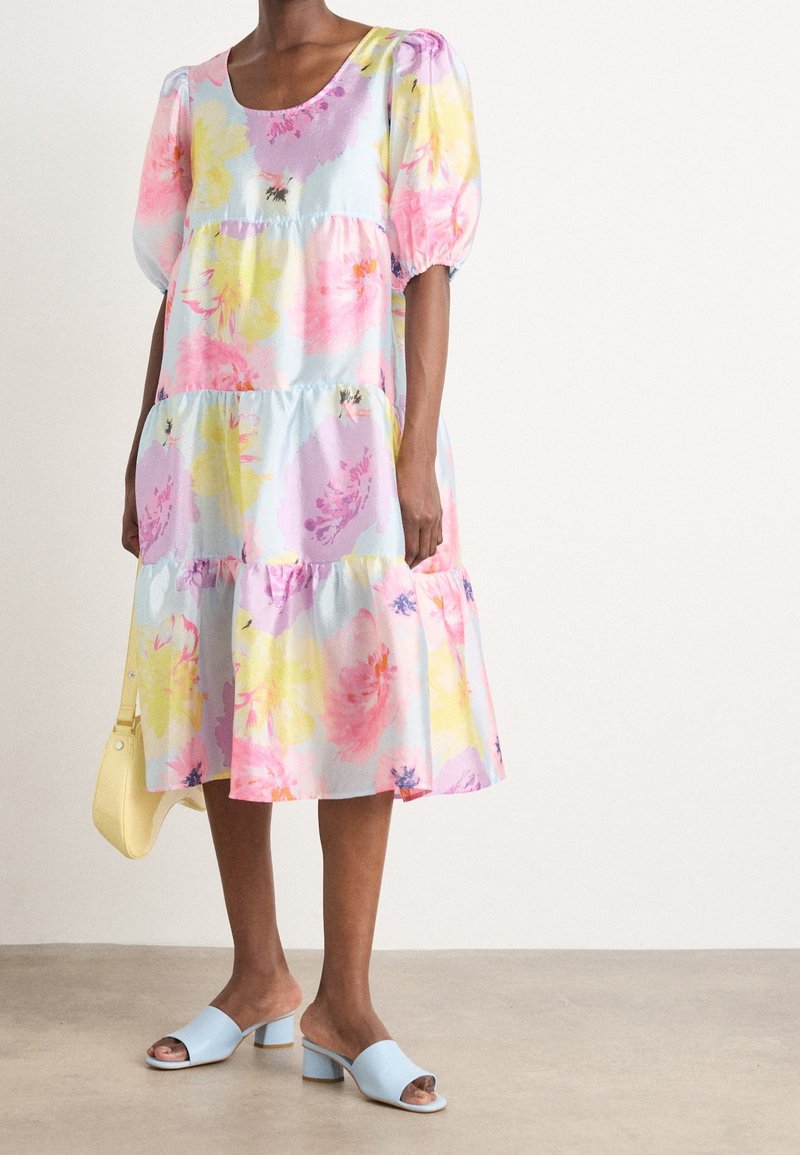 Femme portant une robe midi à volants florale pastel, des sandales à talons bleu clair, et tenant un petit sac à main jaune.