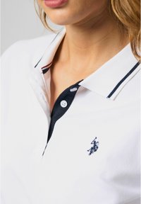 Witte polo met een marineblauwe kraag en knoopsluiting, met rode en witte strepen. Bevat een klein zwart logo op de borst.