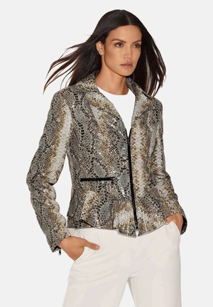 Femme aux longs cheveux foncés porte une veste métallique zippée à motif peau de serpent et un pantalon blanc, mains posées de manière décontractée dans les poches, fond neutre.