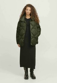 Gepolsterte olivgrüne Jacke mit rundem gesteppte Design, mit hohem Kragen und Druckknöpfen, getragen über einem schwarzen langen Kleid und kniehohen Stiefeln.