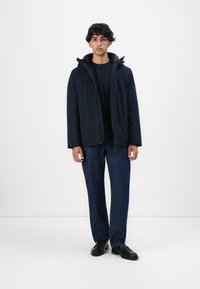 Ung mann som står iført en marineblå jakke, mørkeblå jeans og svarte loafers, med et nøytralt uttrykk mot en enkel hvit bakgrunn.