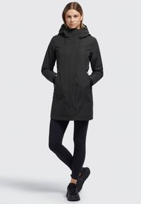 Veste noire imperméable avec un col haut, une capuche et des poches latérales, présentant un design élégant et épuré. Portée sur des leggings noirs et des chaussures.