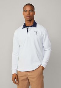 Valkoinen pitkähihainen polo-paita, jossa on tummansininen kaulus. Paidassa on brodeerattu "Hackett London" -logo ja numero 1 yksityiskohtana rinnassa. Yksinkertainen muotoilu.