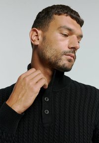 Zwarte gebreide trui met een hoge kraag, voorzien van een ribbelstructuur en twee knopen bij de hals. Close-up van een hand die de kraag aanpast.