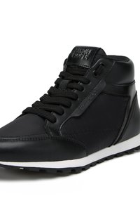 Zapatilla alta negra con una mezcla de materiales lisos y texturizados, con una suela blanca y cordones negros, y un logotipo en relieve en la lengüeta.