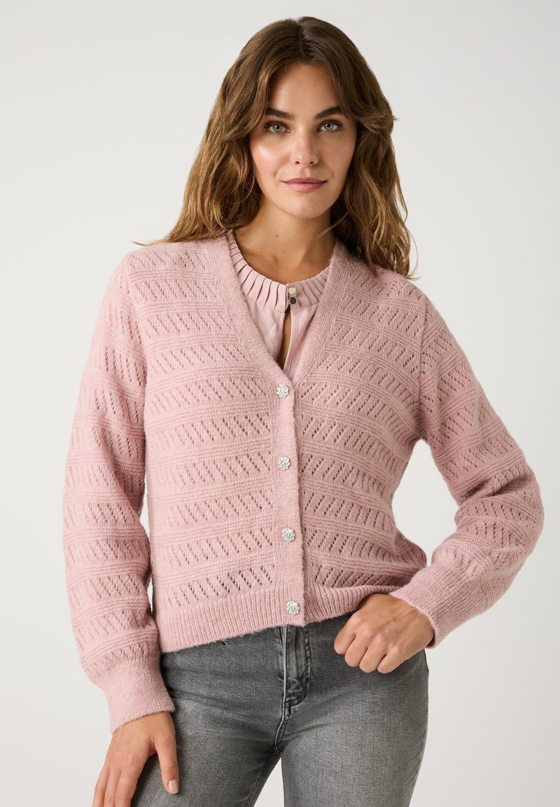 Helles rosa Strickjacke mit V-Ausschnitt und strukturiertem Muster. Verfügt über runde, blumige Knöpfe und lange Ärmel. Kombiniert mit grauen Jeans.