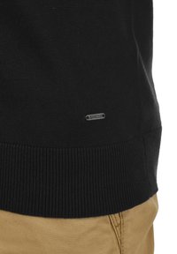 Sort strikket sweater med ribbet kant og et lille sølvmærkat. Mixet med khakifarvede bukser.