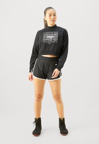 Sweatshirt noir court avec texte graphique blanc, associé à un short noir avec des bordures blanches. Le modèle porte des bottes de sport noires.