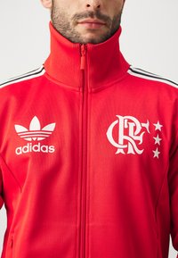 Red zip-up jacket featuring ribbed collar and white accents. Adidas logo on left chest, club emblem with stars on right. Textured fabric.
