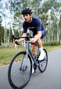 Vélo de route gris avec des pneus fins noirs, freins à disque et une finition mate. Cycliste en maillot bleu marine et chaussures blanches, roulant sur un chemin pavé.