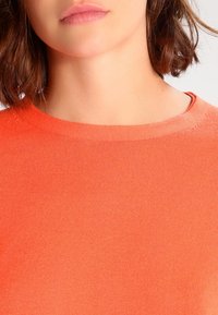 Pull tricoté orange avec un col rond, une texture douce et une finition côtelée. Le tissu semble léger et lisse.