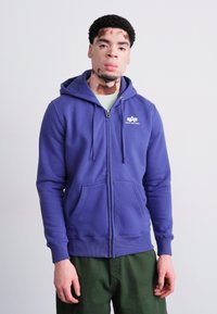 Fialová mikina na zips z mäkkej tkaniny, s klokankovou vreckom a kapucňou na šnúrku. Biele logo na hrudi.