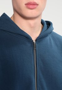 Bluza z kapturem w kolorze ciemnoniebieskim, wykonana z bawełny, z całkowitym zapięciem na zamek błyskawiczny w kolorze srebrnym. Posiada teksturowany materiał oraz usztywniony kaptur.