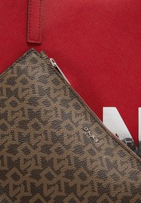 Brun dragkedjepåse med mönstrad textur, prydd med en dragkedja i silver och en DKNY-logotyp, mot en röd canvasbakgrund.