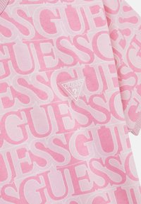Rosa bomullströja med ett upprepat "GUESS"-logotyp i olika nyanser av rosa, med en liten triangulär varumärkespatch nära fållen.