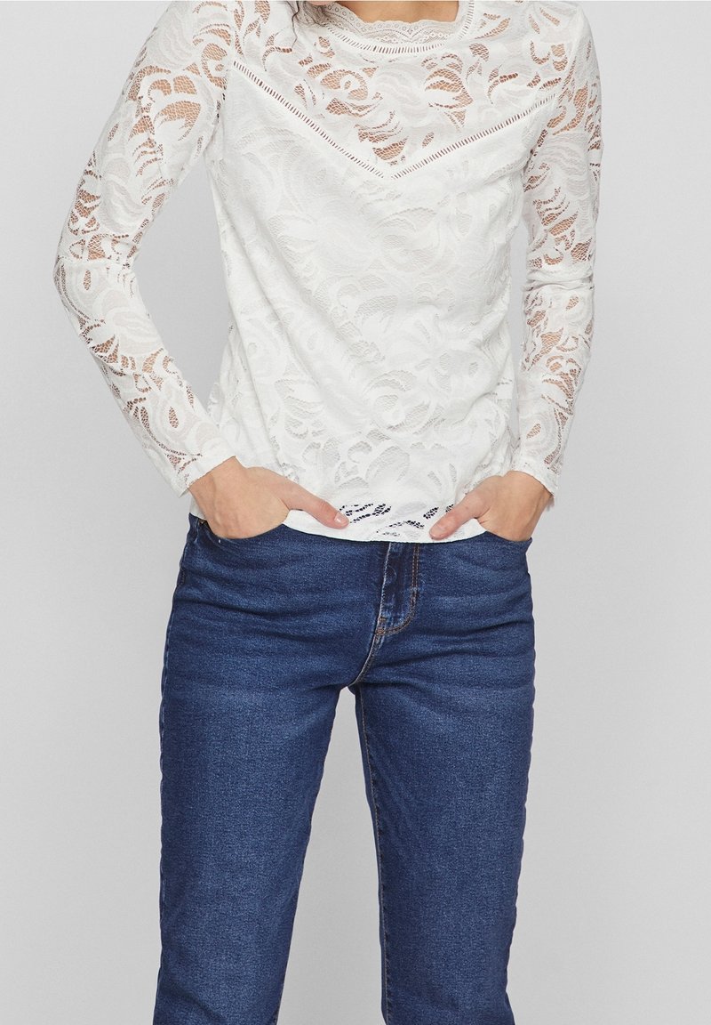 Vit spets tröja med lång ärm som har ett blommönster och ribbade detaljer vid halsringningen, bärs ihop med mörkblå jeans.