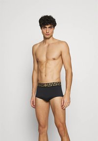 Sorte badeshorts med et gullmønstrete midjebånd og emblem, laget av glatt stoff med en tettsittende design.