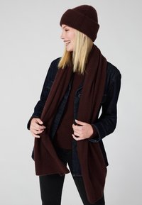 Bordeaux gebreide muts en bijpassende sjaal gedragen over een donkere denimjas en trui. De sjaal hangt naar beneden en creëert een gelaagd effect.