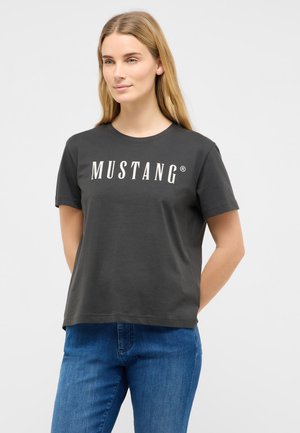 Junge Frau mit langen blonden Haaren, die ein schwarzes "MUSTANG"-T-Shirt und blaue Jeans trägt, steht mit den Händen hinter dem Rücken, heller Hintergrund.