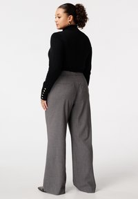 Vrouw met krullend haar in een zwarte coltrui en grijze wijde broek, staand en kijkend naar links tegen een effen witte achtergrond.