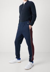 Mörkblå zip-up tröja med texturerad yta kombinerad med mörkblå joggingbyxor med en vinröd sidopanel och vit kantning. Vita sneakers.