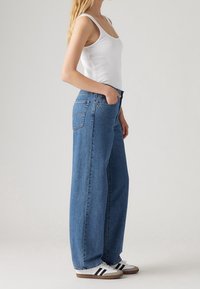 High-waisted, wide-leg blue jeans in denim with a classic five-pocket design and a subtle back pocket detail, worn with sporty shoes.