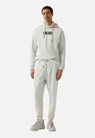 Jonge man staand, gekleed in een lichtgrijze hoodie met de tekst "FIRE&ICE", bijpassende joggingsbroek en grijze sneakers met oranje accenten.
