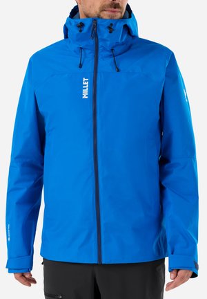 Homme portant une veste imperméable bleu vif avec capuche, fermeture éclair à l'avant et logo sur la poitrine et la manche, pantalon noir visible de la taille vers le bas.