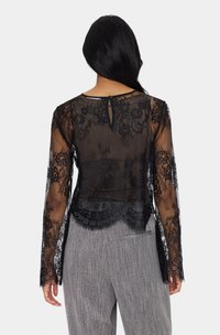 Fekete csipkeblúz hosszú ujjal, áttetsző anyagból, virágmintával, hullámos szegéllyel és hátsó kulcslyuk részlettel, szürke csíkozott nadrággal párosítva.
