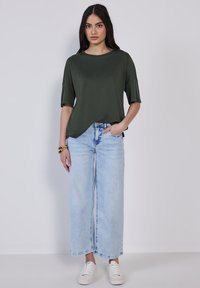 Hnědozelený top s krátkým rukávem v uvolněném střihu, spárovaný s světle modrými širokými džíny a bílými teniskami. Jednoduchá, hladká tkanina bez viditelných vzorů.