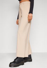 Pantalons larges beige en tissu lisse, dotés d'une fermeture à bouton à l'avant et de poches latérales, associés à des bottes noires à lacets.