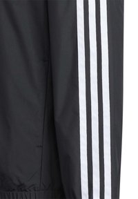Chubasquero negro con tres franjas blancas a lo largo del costado, textura suave y puños elásticos con un ajuste relajado.