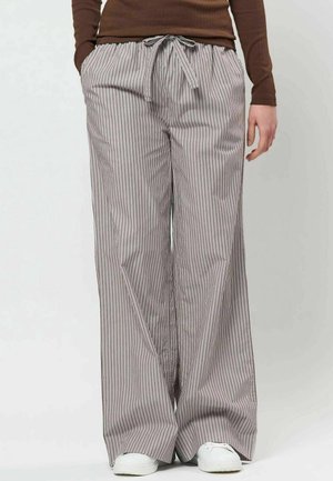 Personne portant un pantalon ample rayé à cordon avec poches, un haut marron à manches longues et des baskets blanches, debout devant un fond uni.