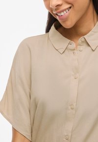 Beige skjorta med krage i lätt tyg, med knäppning fram och korta ärmar. Slen yta med subtil lyster.