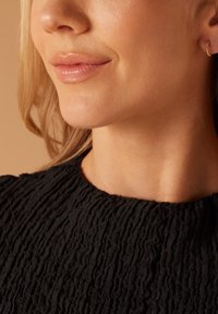 Vêtement noir texturé avec un col haut et froncé, porté près d'un teint clair. Des détails de boucles d'oreilles en or sont visibles.
